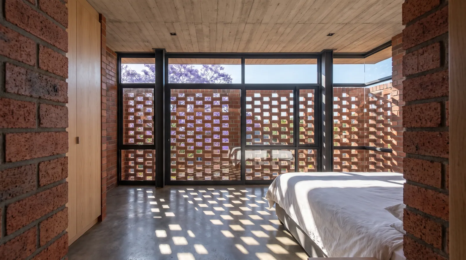 A bedroom with full-height glass and a hit-and-miss brick screen outside it; winter sun throws a chequered pattern of light across the concrete floor and the bed.