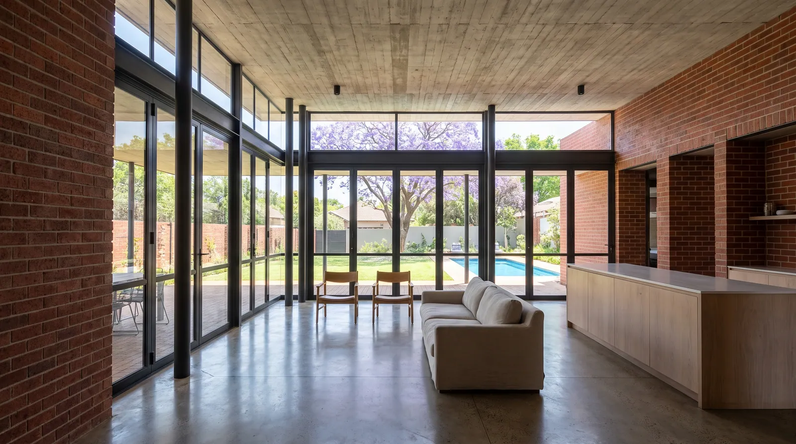 The living room looking north through a wall of steel-framed glass to the shaded stoep, the pool and a jacaranda; a concrete ceiling, a brick wall and a timber kitchen island.